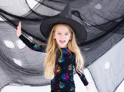Child wearing a Halloween-themed outfit with colorful pumpkins and a witch hat against a spider web background.