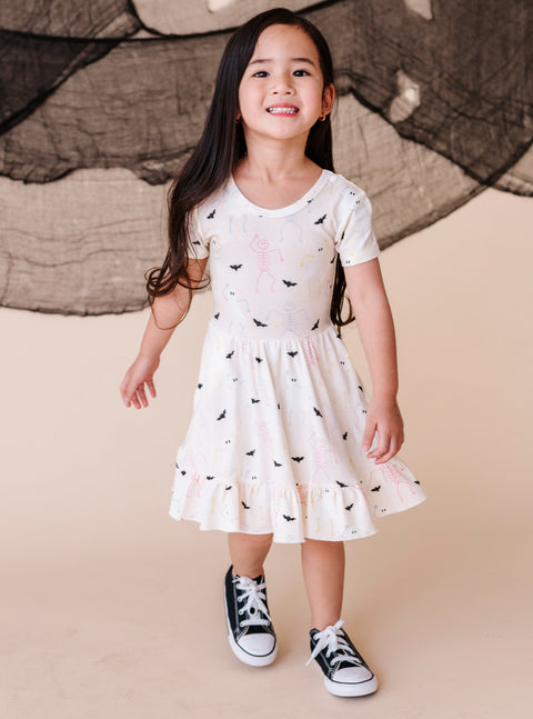 Young girl wearing a cream colored  dress with halloween pattern and black sneakers against a beige background