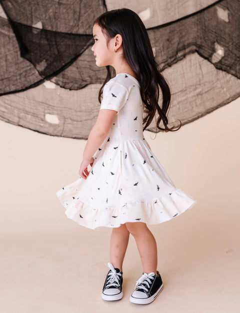 Young girl in a cream dress with black patterns and black sneakers standing against a neutral background.