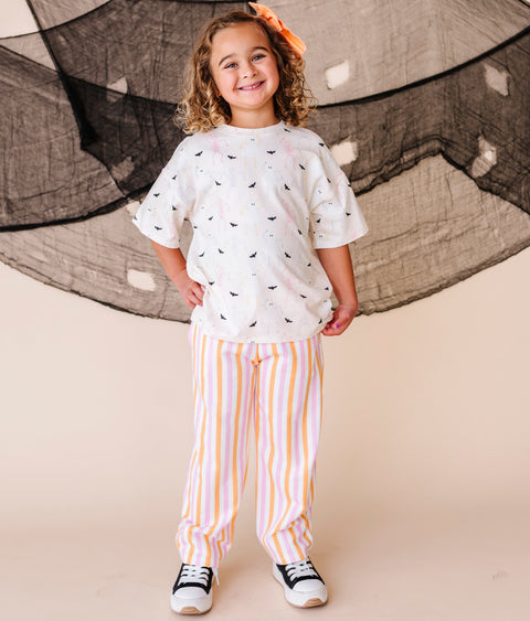 Child wearing a cream shirt with halloween patterns and striped pants, standing in front of large netting.