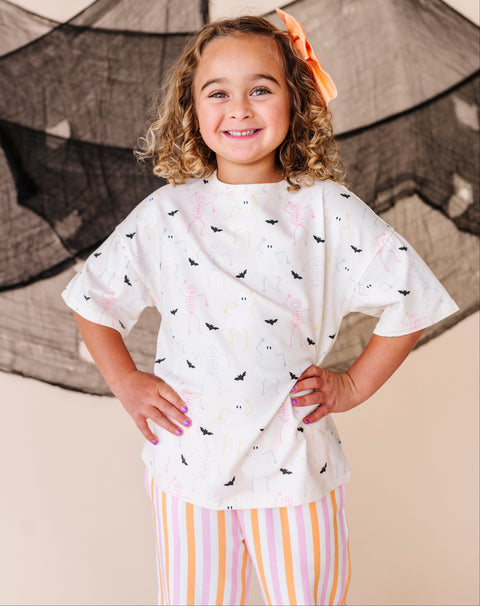Young girl wearing a cream shirt with halloween designs and striped pants, standing in front of a net.
