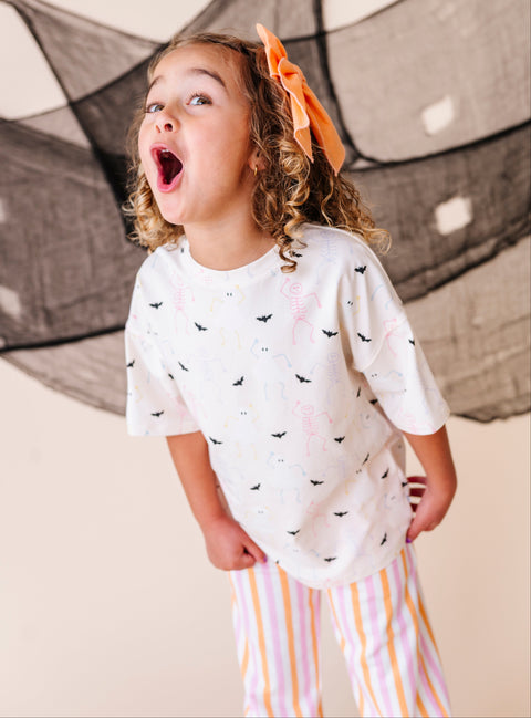 Child wearing a cream shirt with halloween patterns and striped pants, standing in front of a black net.