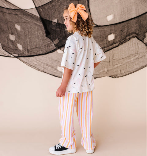 Child wearing a cream shirt with halloween themed patterns and striped pants, standing in front of large netting.