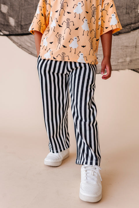 Child wearing a Halloween-themed shirt with skeletons and ghosts, black and white striped pants, and white sneakers on a beige background.