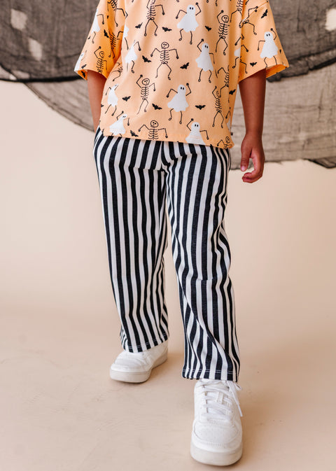 Child wearing a Halloween-themed shirt with skeletons and ghosts, black and white striped pants, and white sneakers on a beige background.