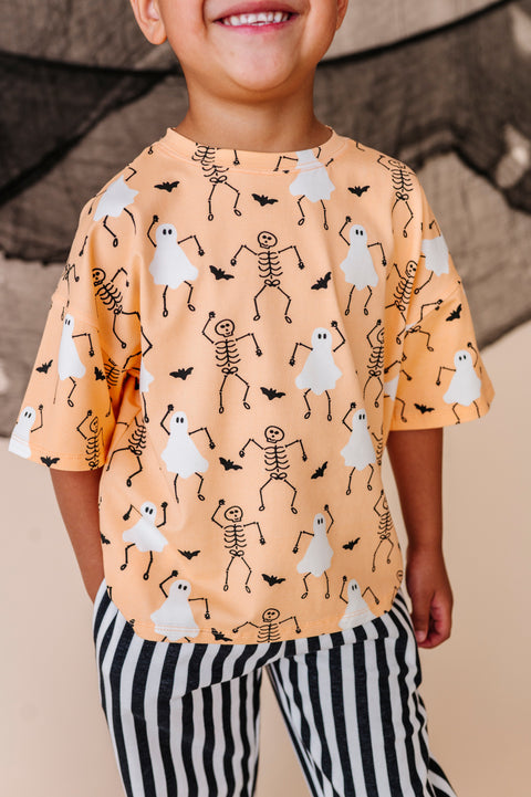 Child wearing a Halloween-themed shirt with ghosts and skeletons on an orange background.