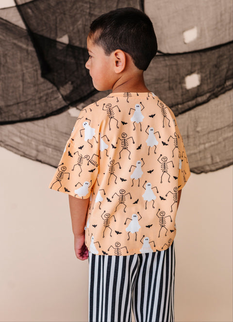 Child wearing a Halloween-themed shirt with ghosts and skeletons in front of a textured wall.