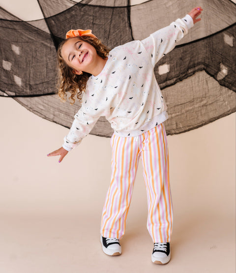 Child wearing a halloween patterned long-sleeve sweatshirt and striped pants standing against a beige background with decorative netting.
