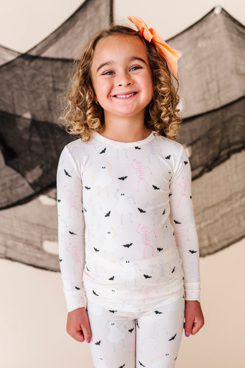 Young girl wearing cream long-sleeve pajamas with halloween patterns and white pants, standing in front of a decorative net.