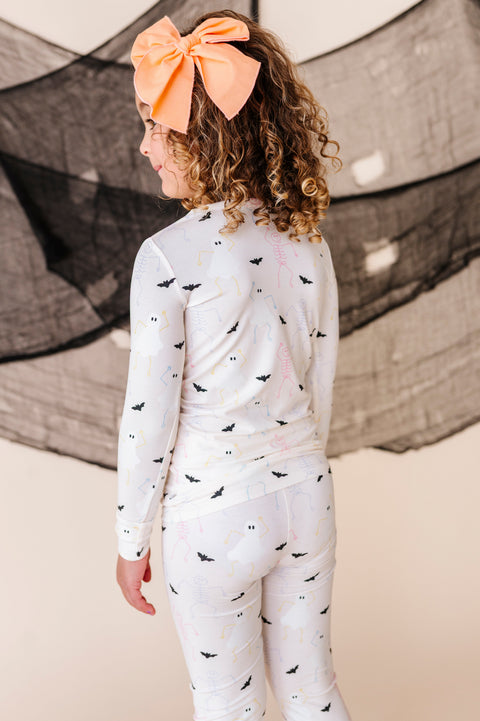 Child wearing a cream pajama set with halloween patterns and an orange bow, standing against a textured wall.