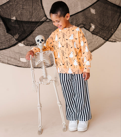 Child in Halloween-themed outfit with skeleton and ghost pattern, standing next to a skeleton decoration.