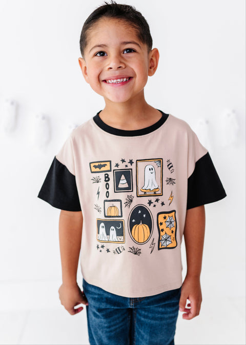 A smiling young boy wearing a light beige t-shirt with black sleeves and a colorful Halloween graphic print, paired with blue jeans, standing against a white wall.