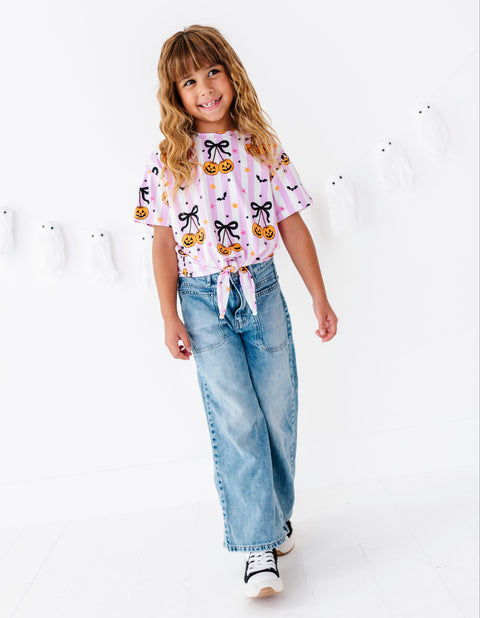 A young girl with blonde hair is smiling and posing in a white studio setting, wearing a light pink short-sleeve t-shirt with a pumpkin and bow pattern, light-wash wide-leg jeans, and white sneakers.