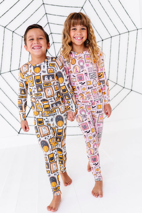 Two children standing barefoot in front of a white studio backdrop with a black spiderweb graphic. The child on the left wears a long-sleeve one-piece pajama with a black, yellow, and gray graphic print, and the child on the right wears a matching long-sleeve one-piece pajama with a pink, white, and orange graphic print.