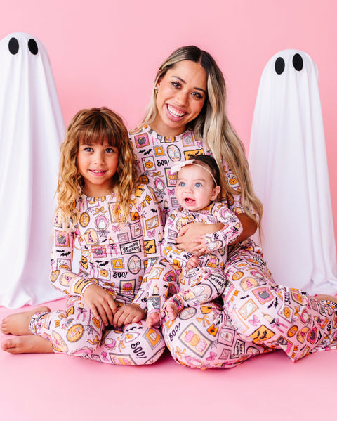 A woman and two children wearing matching pink patterned pajamas sit together on a pink studio floor, flanked by two white ghost props with black eyes. The pajamas have a colorful food and snack collage print.