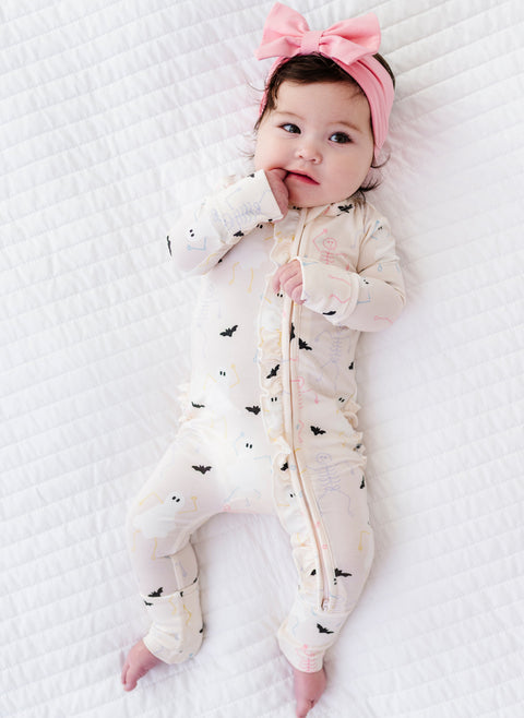 Baby lying on a white textured surface wearing a white onesie with black bat and halloween patterns and a pink headband.