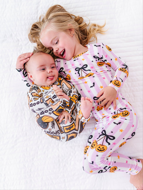 Two children lying on a white quilted bed wearing matching Halloween-themed long-sleeve pajamas. The older child has blonde hair and wears a light pink pajama set with black bows, orange pumpkins, and small stars.