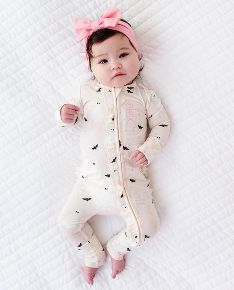 Baby wearing a cream onesie with bat and Halloween patterns and a pink headband on a white textured surface.