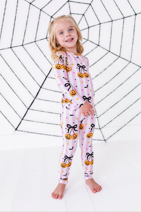 A young girl stands barefoot in front of a white wall with a large black spiderweb print, wearing a pink long-sleeve pajama set with a Halloween print of jack-o'-lanterns, black bows, and bats.