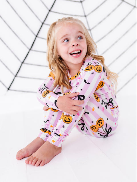 A young girl with blonde hair sits smiling on a white floor, wearing a pink and white striped pajama set with a Halloween pumpkin and spider print. A black spiderweb graphic is printed on the wall behind her.